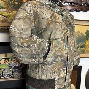 Puede incluir: Chaqueta Carhartt con estampado de camuflaje, capucha y dobladillo acanalado marr&oacute;n. La chaqueta presenta cierre de cremallera completa, dos bolsillos delanteros y un parche con el logo de Carhartt. La chaqueta se exhibe en un maniqu&iacute;.