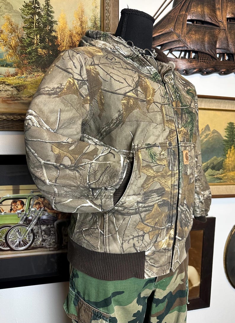 Puede incluir: Chaqueta Carhartt con estampado de camuflaje, capucha y dobladillo acanalado marr&oacute;n. La chaqueta presenta cierre de cremallera completa, dos bolsillos delanteros y un parche con el logo de Carhartt. La chaqueta se exhibe en un maniqu&iacute;.