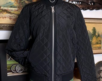 May include: Black quilted bomber jacket with a silver zip. The jacket features a diamond pattern and ribbed collar and cuffs. The jacket is displayed on a mannequin with a black turtleneck and a silver chain necklace.