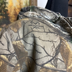 Puede incluir: Primer plano de una chaqueta de camuflaje con el logotipo de Realtree Xtra. La chaqueta presenta un patr&oacute;n de color marr&oacute;n, verde y beige, que imita un entorno forestal. Las costuras son visibles.