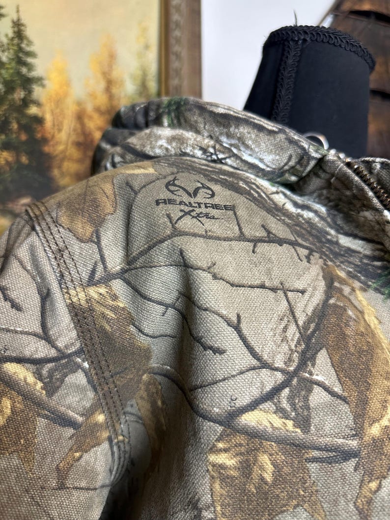 Puede incluir: Primer plano de una chaqueta de camuflaje con el logotipo de Realtree Xtra. La chaqueta presenta un patr&oacute;n de color marr&oacute;n, verde y beige, que imita un entorno forestal. Las costuras son visibles.