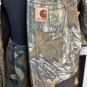 Puede incluir: Una chaqueta de camuflaje Carhartt y pantalones a juego. La chaqueta presenta un parche de cuero marr&oacute;n con el logotipo de Carhartt y un logotipo de Realtree. La chaqueta tiene cierre de cremallera y un dobladillo acanalado marr&oacute;n. Los pantalones tienen cierre de bot&oacute;n.