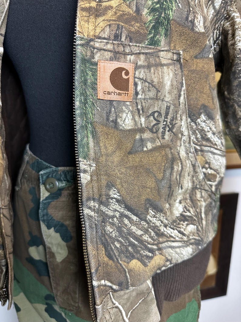 Puede incluir: Una chaqueta de camuflaje Carhartt y pantalones a juego. La chaqueta presenta un parche de cuero marr&oacute;n con el logotipo de Carhartt y un logotipo de Realtree. La chaqueta tiene cierre de cremallera y un dobladillo acanalado marr&oacute;n. Los pantalones tienen cierre de bot&oacute;n.