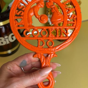 May include: Orange cast iron trivet with the words "Kissin' Don't Last, Cookin' Do" and a couple design. The trivet has a decorative handle. A kitchen decor item.