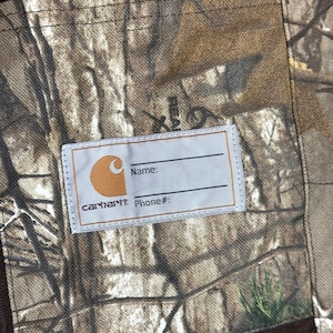 Puede incluir: Primer plano de una tela de camuflaje Carhartt con una etiqueta para el nombre y el n&uacute;mero de tel&eacute;fono. La etiqueta es blanca con el logotipo y el texto de Carhartt. La tela tiene un patr&oacute;n marr&oacute;n y verde.