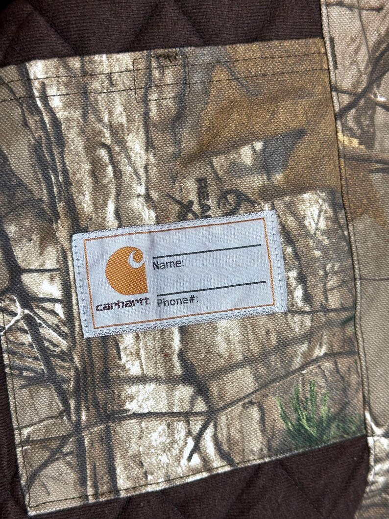 Puede incluir: Primer plano de una tela de camuflaje Carhartt con una etiqueta para el nombre y el n&uacute;mero de tel&eacute;fono. La etiqueta es blanca con el logotipo y el texto de Carhartt. La tela tiene un patr&oacute;n marr&oacute;n y verde.