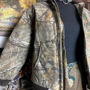 Puede incluir: Una chaqueta de camuflaje con capucha, cierre de cremallera y bolsillos delanteros. La chaqueta se exhibe en un maniqu&iacute; negro. La paleta de colores de la chaqueta incluye tonos de marr&oacute;n, verde y beige, con un dobladillo y pu&ntilde;os acanalados marrones.