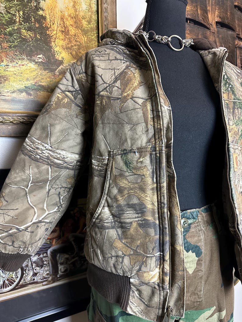 Puede incluir: Una chaqueta de camuflaje con capucha, cierre de cremallera y bolsillos delanteros. La chaqueta se exhibe en un maniqu&iacute; negro. La paleta de colores de la chaqueta incluye tonos de marr&oacute;n, verde y beige, con un dobladillo y pu&ntilde;os acanalados marrones.