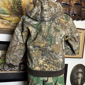 Puede incluir: Chaqueta de camuflaje con capucha y dobladillo y pu&ntilde;os de canal&eacute; marrones. La chaqueta presenta un estampado de bosque en tonos marrones, verdes y beige. Se muestra la parte trasera de la chaqueta, con la capucha puesta.