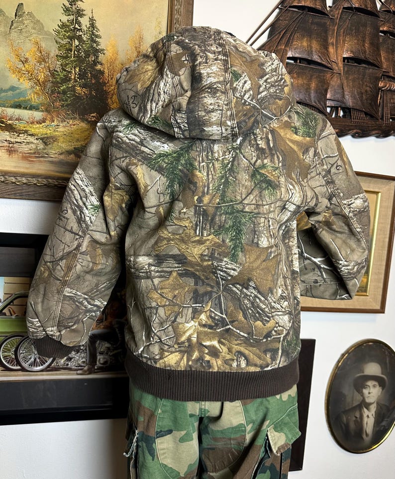 Puede incluir: Chaqueta de camuflaje con capucha y dobladillo y pu&ntilde;os de canal&eacute; marrones. La chaqueta presenta un estampado de bosque en tonos marrones, verdes y beige. Se muestra la parte trasera de la chaqueta, con la capucha puesta.