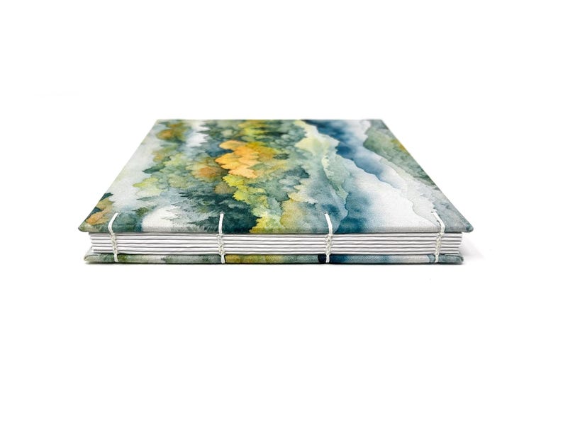 May include: A watercolor landscape journal with a blue, green, and yellow design. The journal is bound with white thread and has a white spine.