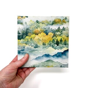 May include: A watercolor painting of a misty forest scene with green and yellow trees and blue mountains in the background.
