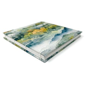 May include: A handmade journal with a watercolor cover featuring a landscape of blue and green mountains with yellow accents. The journal is bound with white stitching.