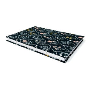 May include: A rectangular, hand-stitched journal with a dark blue floral pattern. The cover features a repeating design of white, pink, and yellow flowers and leaves. The pages are bound with white thread, creating a unique and decorative spine.