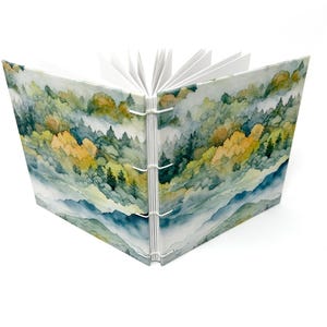 May include: A watercolor-style journal with a cover depicting a misty forest scene with green and yellow trees. The journal is bound with white stitching and has blank pages inside.