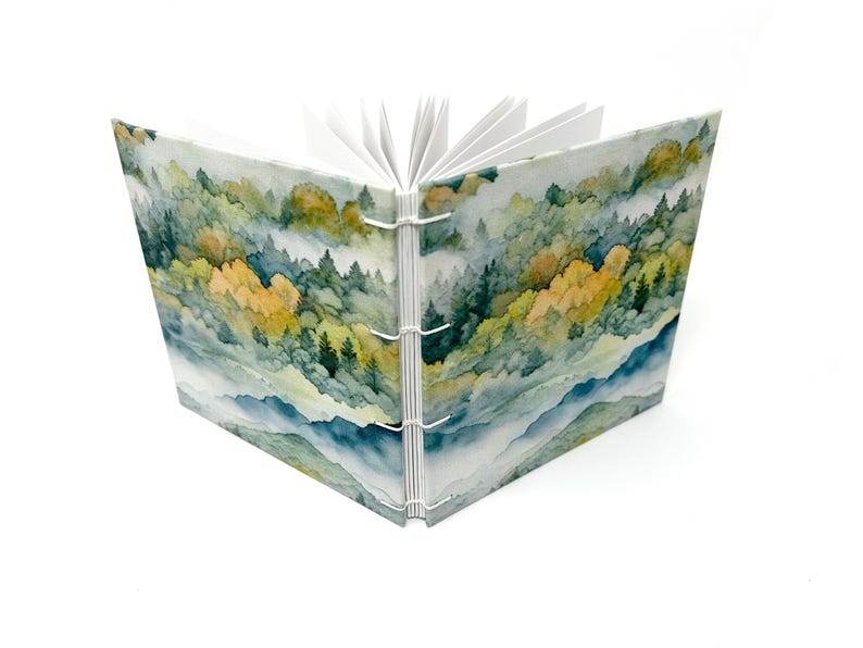 May include: A watercolor-style journal with a cover depicting a misty forest scene with green and yellow trees. The journal is bound with white stitching and has blank pages inside.