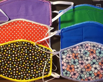 May include: Six fabric face masks with different patterns. The masks are all made of cotton and have elastic ear loops. The patterns include polka dots, flowers, and solid colors. The masks are all in a stack, with the top mask being purple, the second mask being green, the third mask being blue, the fourth mask being red with white polka dots, the fifth mask being black with yellow and pink polka dots, and the sixth mask being white with pink, blue, and green flowers.