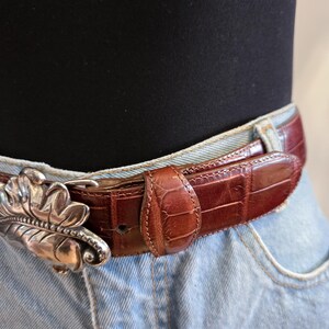 Peut inclure: Une ceinture en cuir marron avec une boucle argent&eacute;e en forme de feuille. La ceinture pr&eacute;sente un motif textur&eacute; en forme de crocodile. La boucle est une conception de feuille d&eacute;corative d&eacute;taill&eacute;e. La ceinture est port&eacute;e avec un jean bleu.