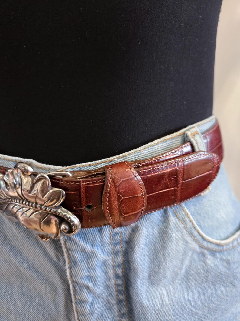 Peut inclure: Une ceinture en cuir marron avec une boucle argent&eacute;e en forme de feuille. La ceinture pr&eacute;sente un motif textur&eacute; en forme de crocodile. La boucle est une conception de feuille d&eacute;corative d&eacute;taill&eacute;e. La ceinture est port&eacute;e avec un jean bleu.