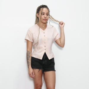 May include: A light pink button-up blouse with short sleeves. The blouse has a fitted bodice and a slightly flared hem.