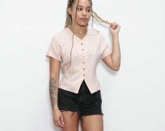 Puede incluir: Una blusa rosa claro con botones y mangas cortas. La blusa tiene un corpiño ajustado y un dobladillo ligeramente acampanado.