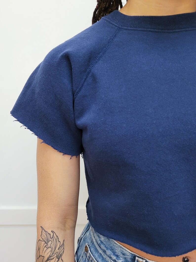 May include: A navy blue cropped sweatshirt with a crew neck and raglan sleeves. The sweatshirt has a distressed hem.