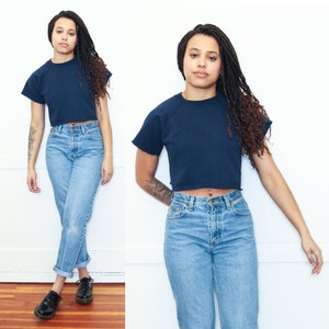 May include: A woman is wearing a navy blue cropped sweatshirt with a short sleeve and a pair of light blue high-waisted jeans.
