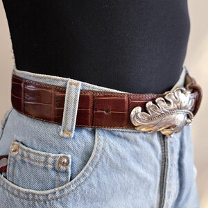 Peut inclure: Une ceinture en cuir marron avec une texture crocodile et une boucle argent&eacute;e en forme de feuille. La ceinture est port&eacute;e avec un jean en denim bleu clair et un haut noir.
