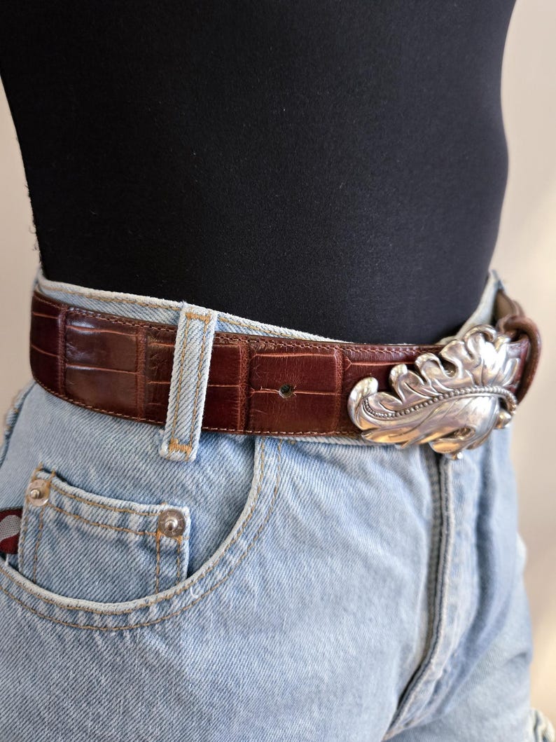 Peut inclure: Une ceinture en cuir marron avec une texture crocodile et une boucle argent&eacute;e en forme de feuille. La ceinture est port&eacute;e avec un jean en denim bleu clair et un haut noir.