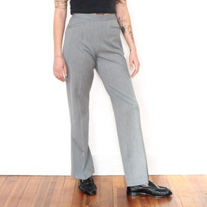 May include: High-waisted gray trousers featuring a straight-leg cut, a clean waistband, and subtle side pockets. The pants are crafted from a smooth, solid-colored fabric and are paired with black leather loafers, creating a classic and neutral look.