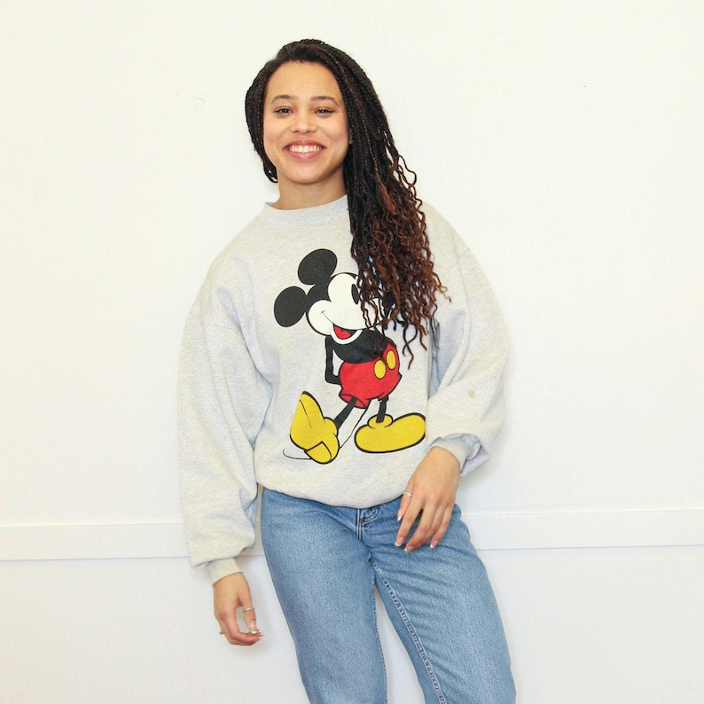 May include: A gray sweatshirt with a large Mickey Mouse graphic on the front. Mickey Mouse is wearing red shorts and yellow shoes. The sweatshirt has long sleeves and a crew neck.