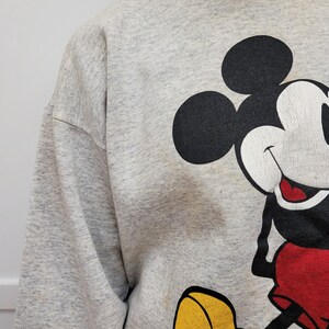 May include: A gray sweatshirt with a large graphic print of Mickey Mouse. Mickey is wearing red shorts and yellow shoes.