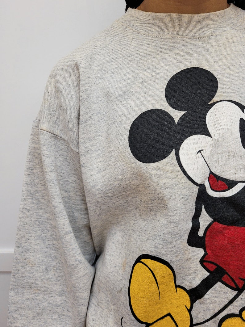 May include: A gray sweatshirt with a large graphic print of Mickey Mouse. Mickey is wearing red shorts and yellow shoes.