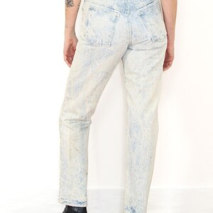May include: High-waisted acid wash denim jeans in a light blue and white mottled pattern. The jeans feature a classic five-pocket design with a tan leather patch on the back waistband, belt loops, and a straight leg cut that reaches the ankles.