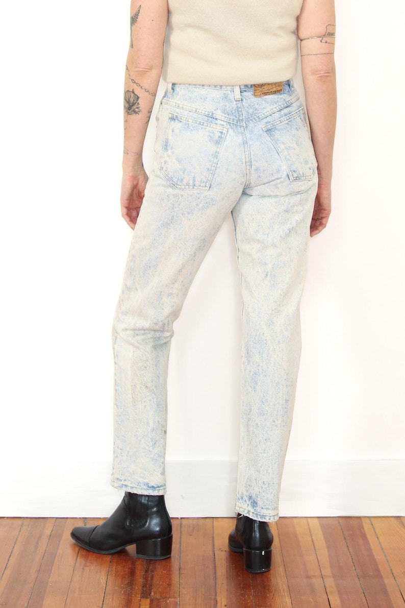 May include: High-waisted acid wash denim jeans in a light blue and white mottled pattern. The jeans feature a classic five-pocket design with a tan leather patch on the back waistband, belt loops, and a straight leg cut that reaches the ankles.