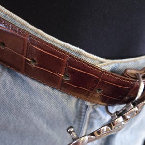 Peut inclure: Une ceinture en cuir marron avec un motif crocodile est attach&eacute;e avec une boucle argent&eacute;e. La ceinture est port&eacute;e avec un jean en denim bleu clair. La boucle a un design d&eacute;coratif.