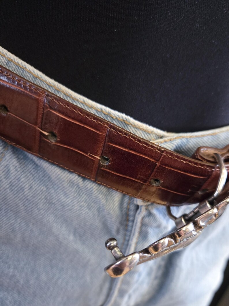 Peut inclure: Une ceinture en cuir marron avec un motif crocodile est attach&eacute;e avec une boucle argent&eacute;e. La ceinture est port&eacute;e avec un jean en denim bleu clair. La boucle a un design d&eacute;coratif.