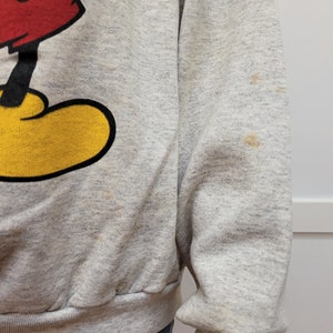May include: A gray sweatshirt with a vintage Mickey Mouse graphic. The sweatshirt has yellow, red, and black details. There are a few light brown stains on the sweatshirt.