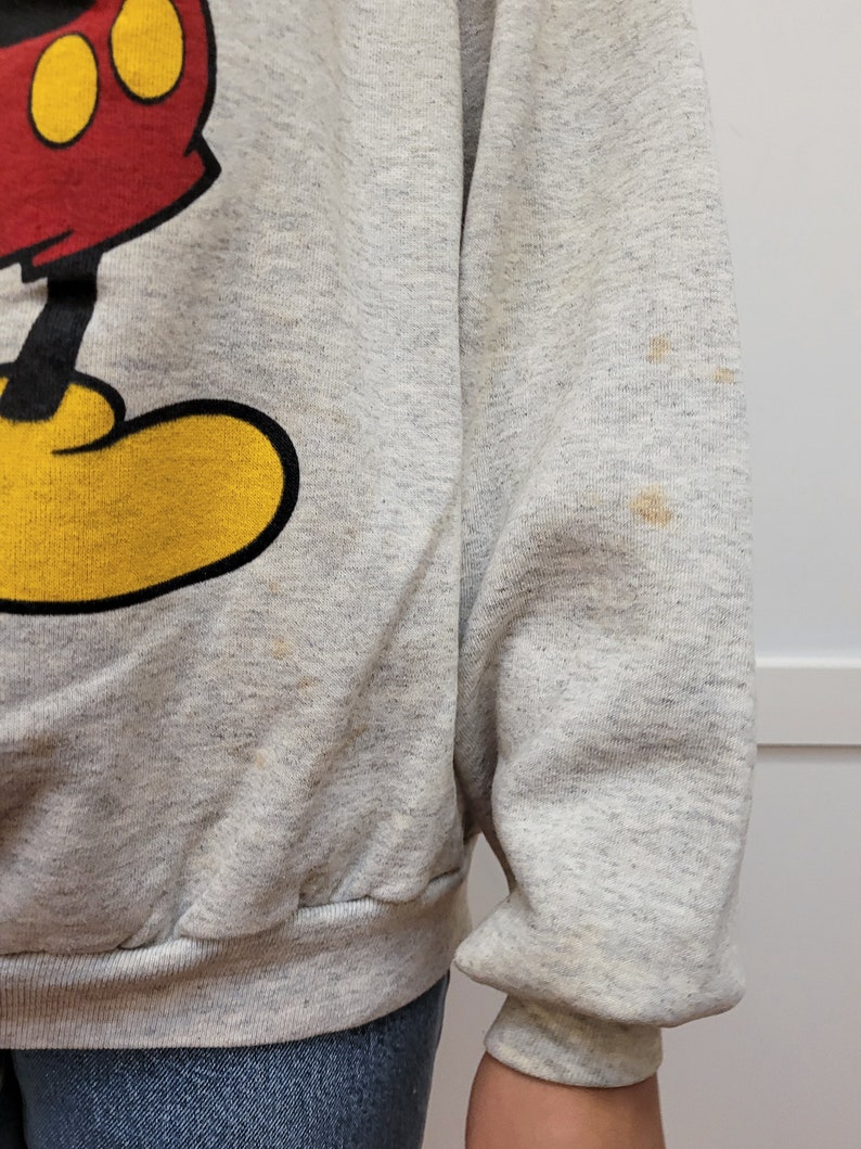 May include: A gray sweatshirt with a vintage Mickey Mouse graphic. The sweatshirt has yellow, red, and black details. There are a few light brown stains on the sweatshirt.