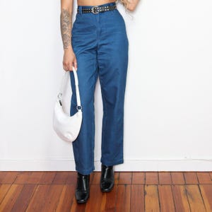 May include: A woman wearing a pair of blue high-waisted jeans with a black studded belt and a white handbag.