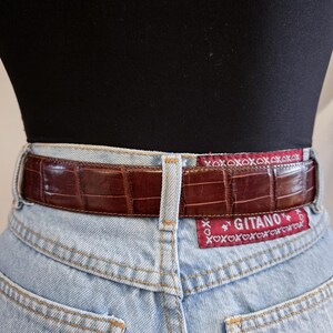 Peut inclure: Une ceinture en cuir marron avec un motif crocodile est port&eacute;e avec un jean en denim bleu clair. La ceinture porte une &eacute;tiquette rouge avec la marque "Gitano" et le texte "xoxoxoxoxox".