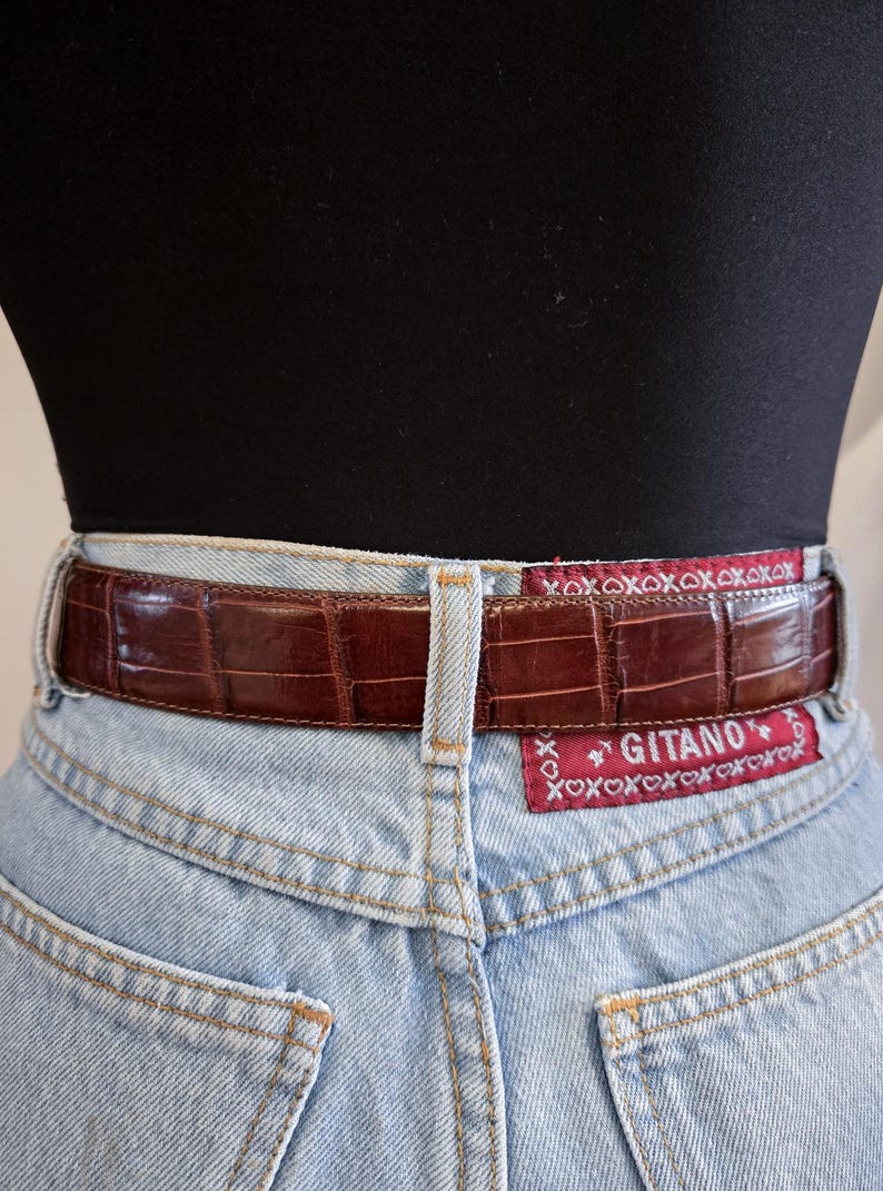 Peut inclure: Une ceinture en cuir marron avec un motif crocodile est port&eacute;e avec un jean en denim bleu clair. La ceinture porte une &eacute;tiquette rouge avec la marque "Gitano" et le texte "xoxoxoxoxox".
