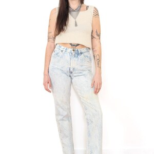 May include: A beige sleeveless ribbed crop top paired with high-waisted acid-wash blue denim jeans and black leather ankle boots. The jeans feature a classic five-pocket design with a button fly closure and a straight-leg cut.