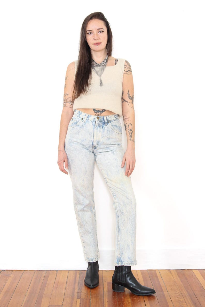May include: A beige sleeveless ribbed crop top paired with high-waisted acid-wash blue denim jeans and black leather ankle boots. The jeans feature a classic five-pocket design with a button fly closure and a straight-leg cut.