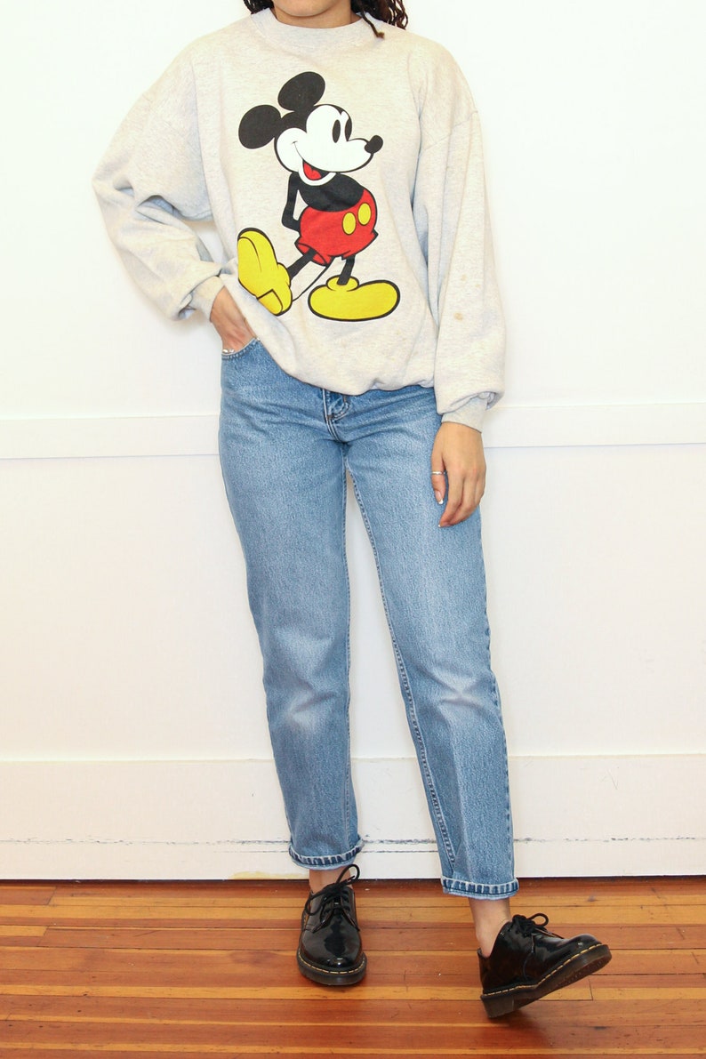 May include: A gray sweatshirt with a Mickey Mouse graphic. The sweatshirt has long sleeves and a crew neck. The person is wearing blue jeans and black shoes.