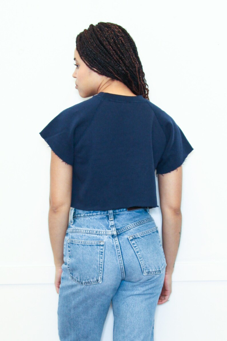 May include: A navy blue cropped sweatshirt with short sleeves and a relaxed fit. The sweatshirt is worn with light blue denim jeans.