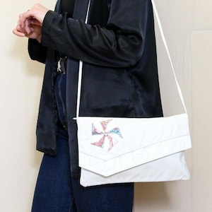 Puede incluir: Un bolso blanco de bandolera con cierre de solapa y un diseño en forma de estrella colorido y estampado en la parte delantera.