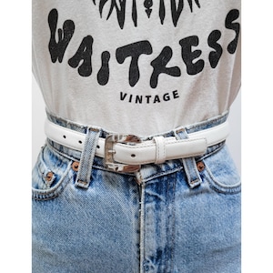 May include: A white leather belt with a silver buckle. The belt is worn over a pair of light blue denim jeans and a white t-shirt with the word "Waitress" printed in black.