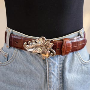Peut inclure: Une ceinture en cuir marron avec une boucle argent&eacute;e en forme de feuille. La ceinture a une texture de peau de crocodile et est port&eacute;e avec un jean en denim bleu clair. La boucle est orn&eacute;e de veines de feuilles complexes et d'un bord d&eacute;coratif.