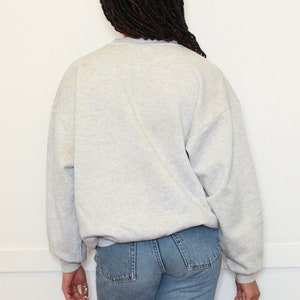 May include: A gray sweatshirt with long sleeves and a crew neck. The sweatshirt is worn by a person with long black braids. The person is wearing blue jeans.